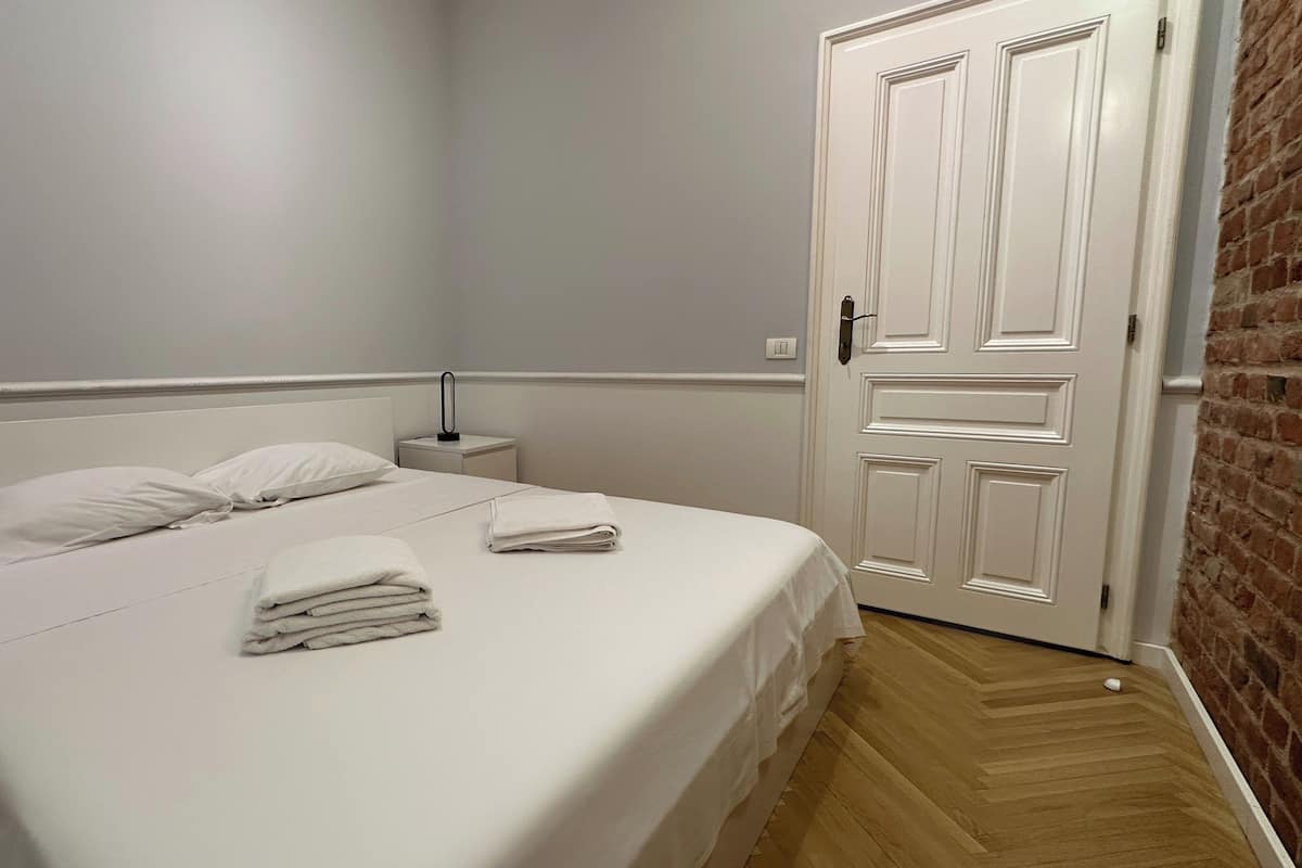 Una cama bien tendida con sábanas y toallas blancas, una mesita de noche con lámpara, y una puerta blanca con cerradura.