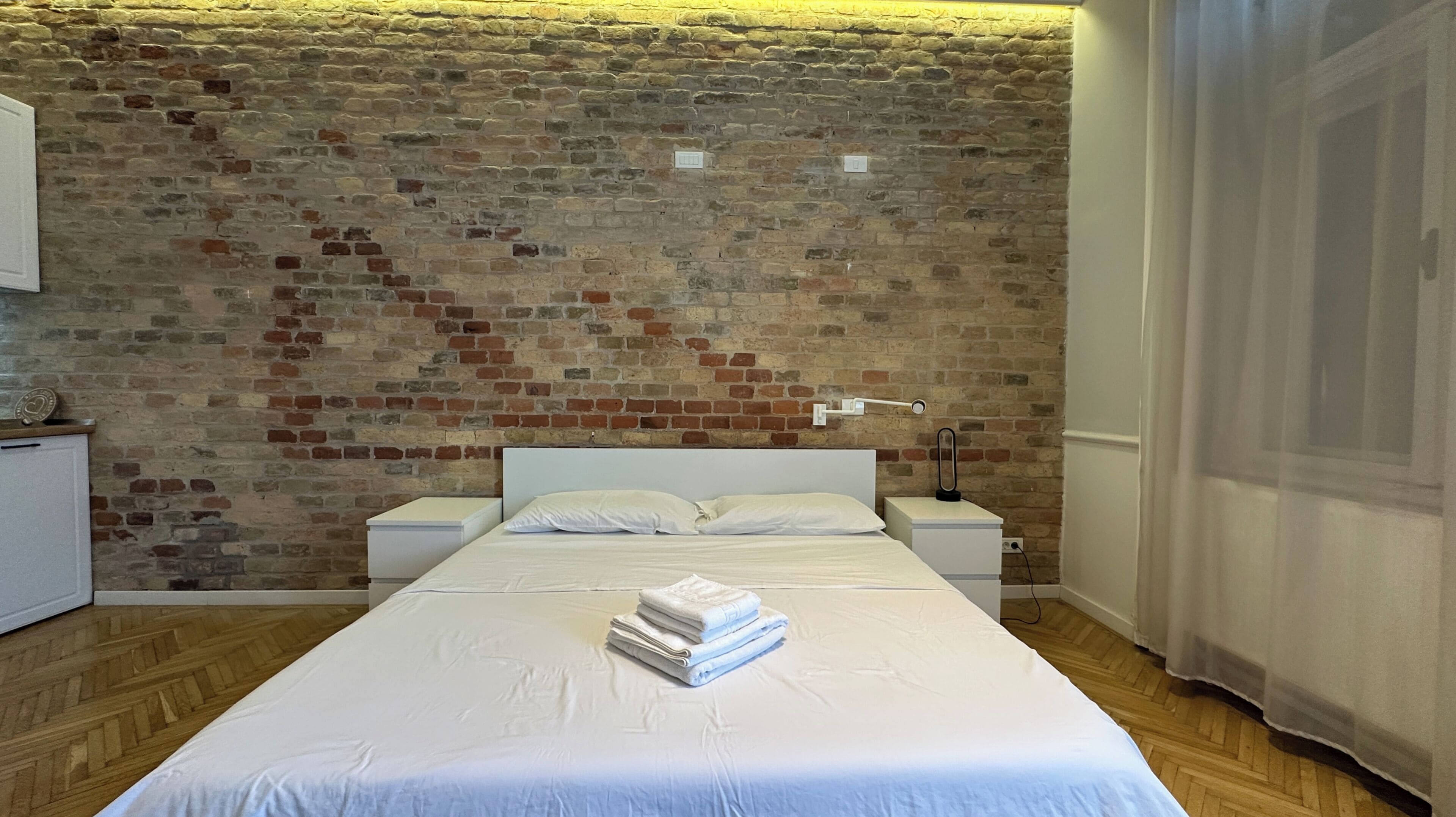 A bedroom with a brick wall, a bed with white bedding, a bedside table, a lamp, and a window with curtains.