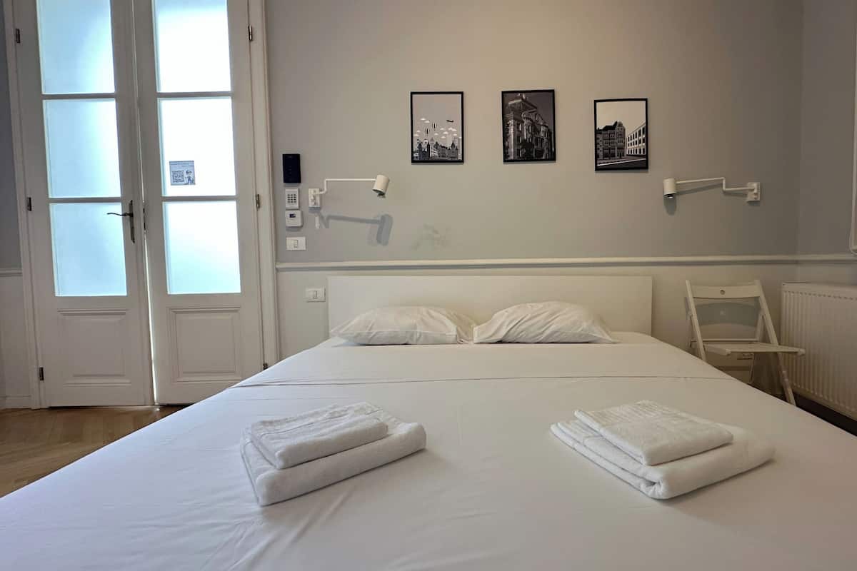 Una habitación de hotel con una cama grande, dos mesitas de noche, un radiador y tres cuadros en la pared.
