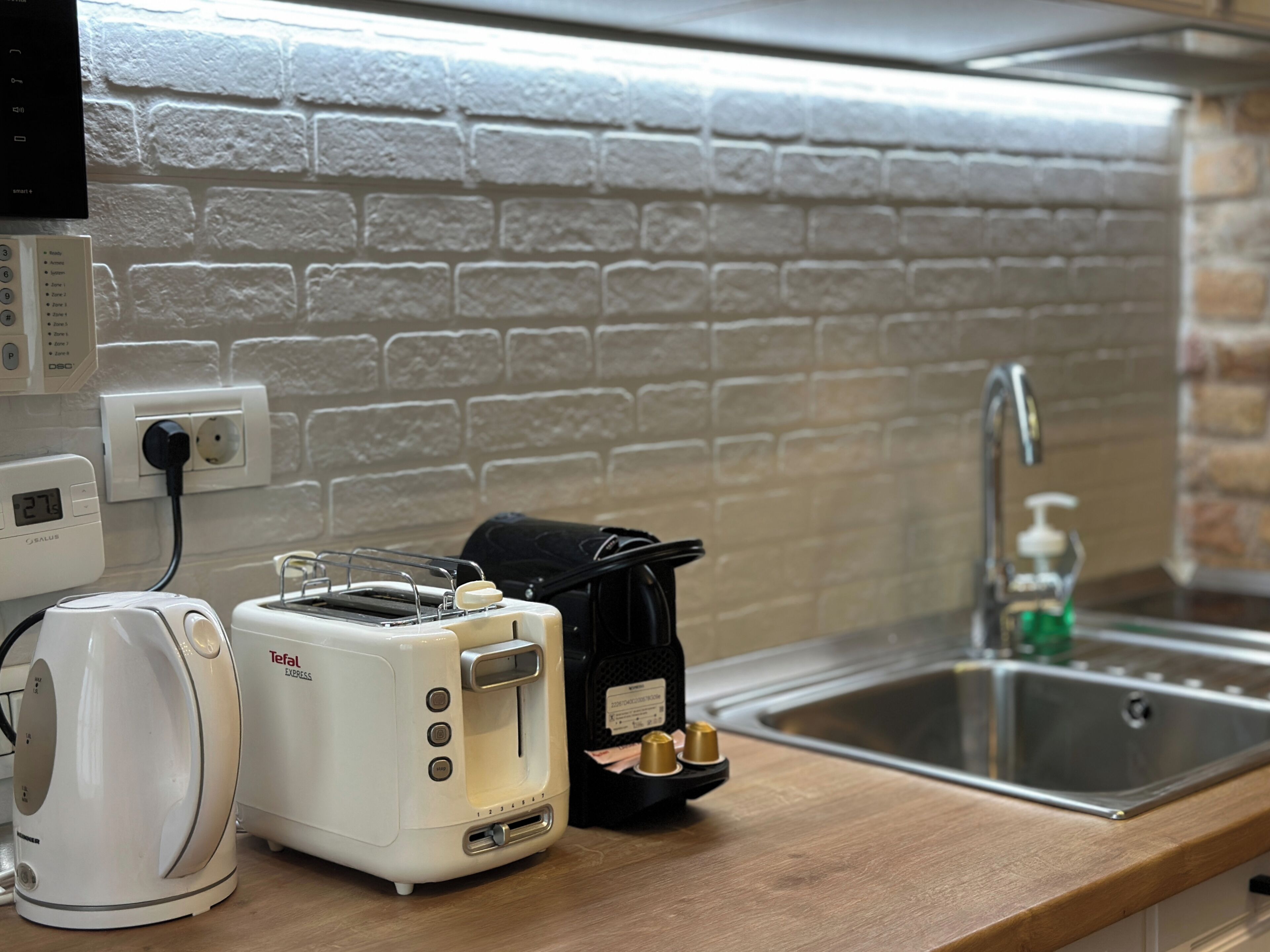 A kitchenette with a white brick wall, a stainless steel sink, a black coffee maker, a white toaster, and a kettle.