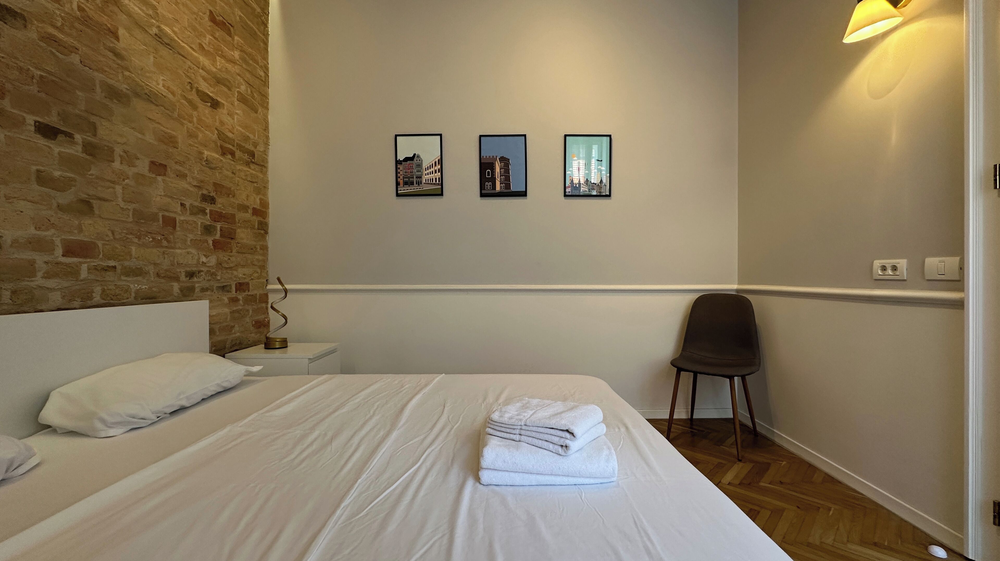 A bedroom with a brick wall, a bed with white linens, a chair, and three framed pictures on the wall.