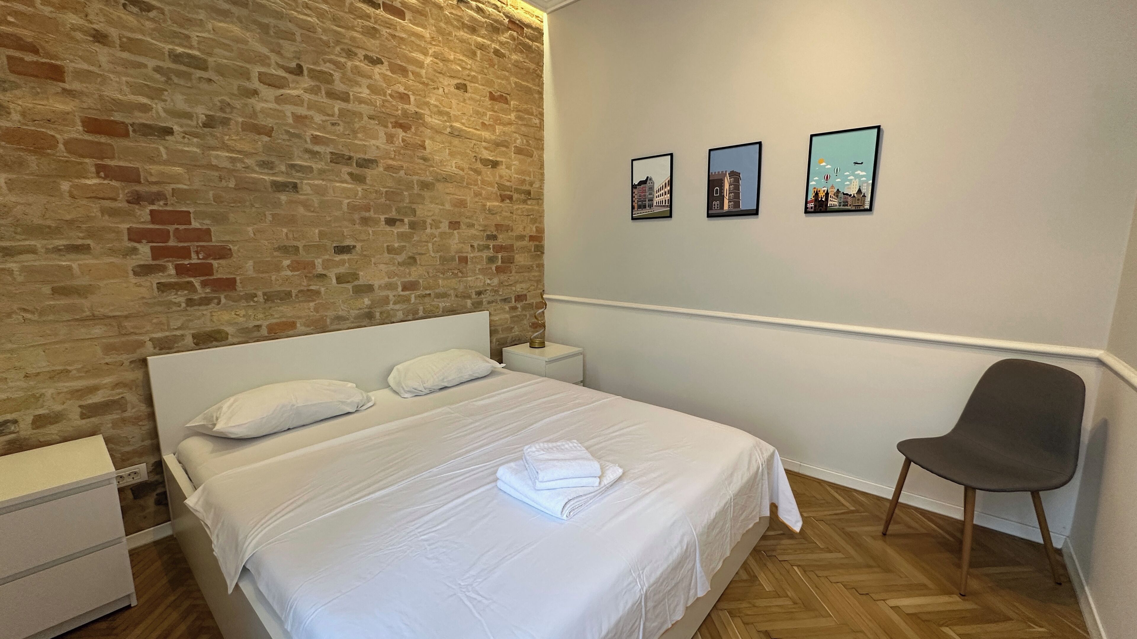 A bedroom with a brick wall, a bed with white linens, a nightstand, a chair, and three framed pictures on the wall.