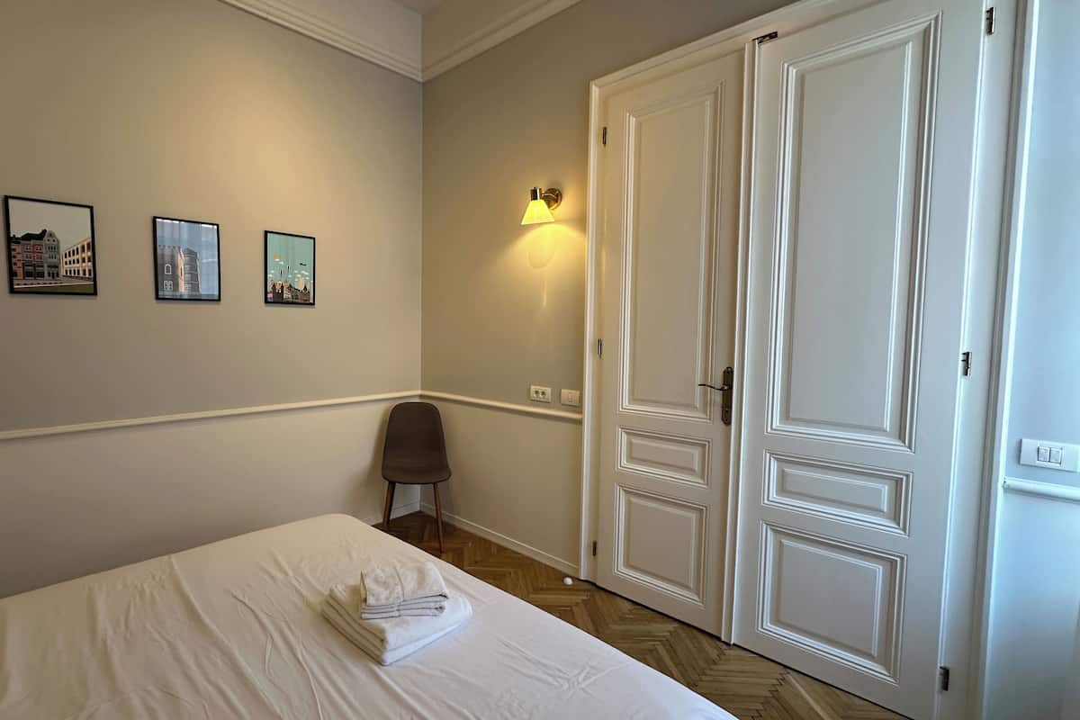 Una habitación de hotel con una cama, una silla, una lámpara fijada a la pared y tres cuadros enmarcados.