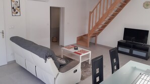 A modern living room with a white sofa, a wooden staircase, a flat-screen TV, and a small table with a red and white pattern. - Maison Chaleureuse au Calme Proximités Commodités (Montaigu-Vendée)