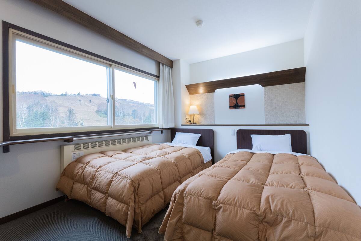 Ein Hotelzimmer mit zwei Betten, einem großen Fenster, einer Heizung und einem kleinen Tisch mit einer Lampe.