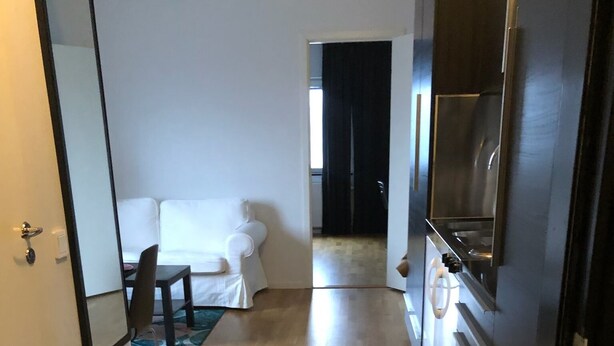 A hotel hallway with a wooden floor, a striped rug, a white sofa, and a kitchen area with a refrigerator. - Årsta Stockholm Aparment 339 (Arsta)
