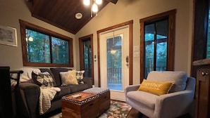 Un salon chaleureux avec un parquet en bois foncé, un canapé gris agrémenté de coussins à motifs, un fauteuil gris clair avec un coussin jaune et une table basse en bois.