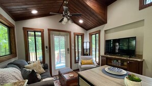 Un salon confortable avec un canapé, une table basse et un téléviseur. Il y a un ventilateur de plafond et une fenêtre donnant sur un paysage verdoyant.