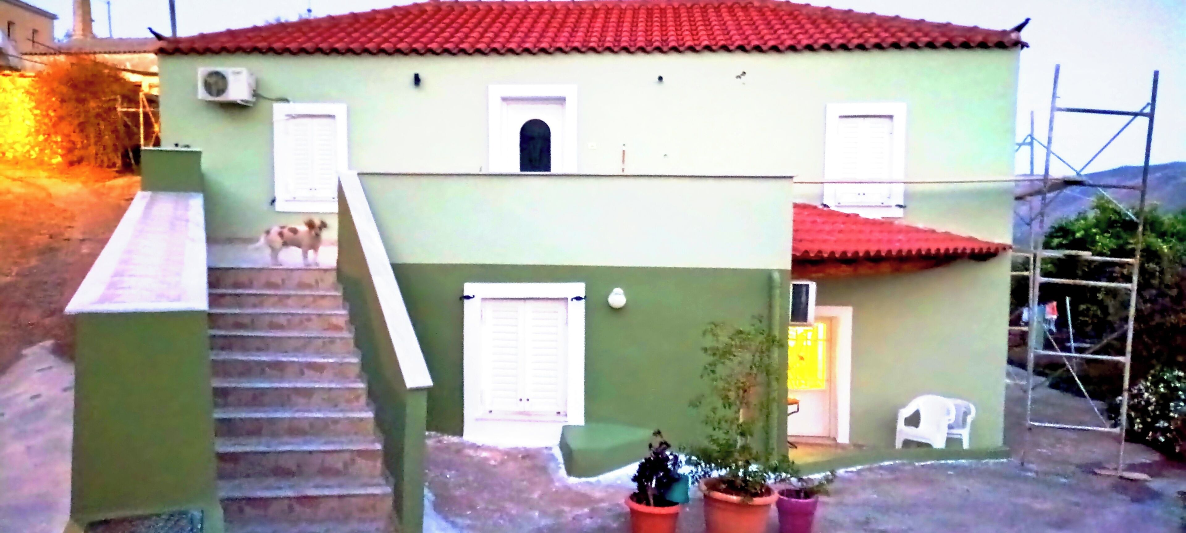 A two-story house with a red-tiled roof, a staircase leading to the upper floor, and a small porch area with potted plants.