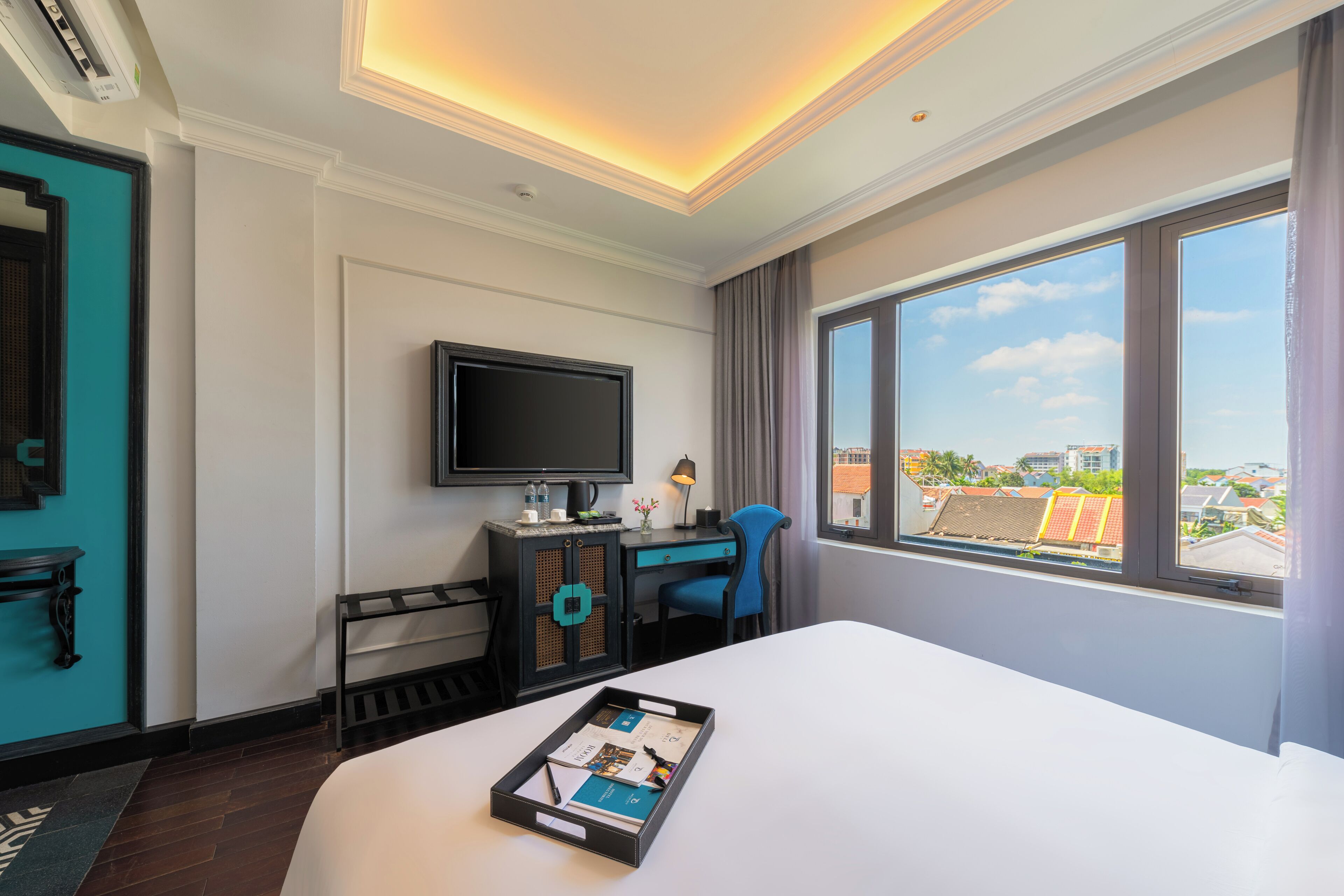 A hotel room with a bed, a desk with a TV, a window with a city view, and a tray with brochures.