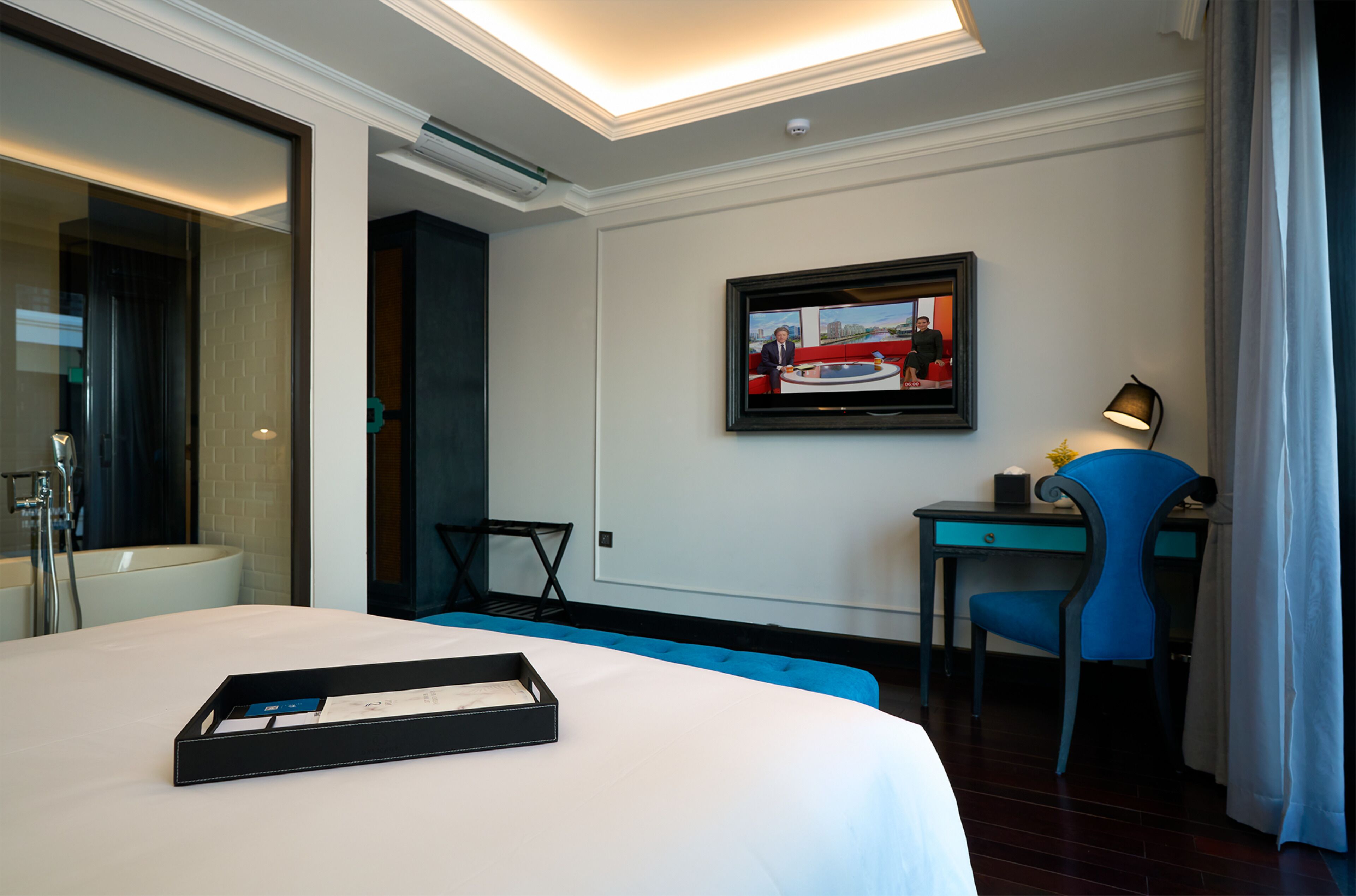 Een hotelkamer met een bed, een bureau, een stoel, een aan de muur bevestigde televisie en een badkamer zichtbaar door een glazen deur.