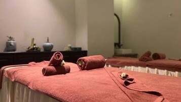 Ein Spa-Raum mit einer Massageliege, die mit einem rosafarbenen Handtuch bedeckt ist, einem Spiegel, der den Raum reflektiert, und einer dunklen Holzkommode mit Dekorationen.