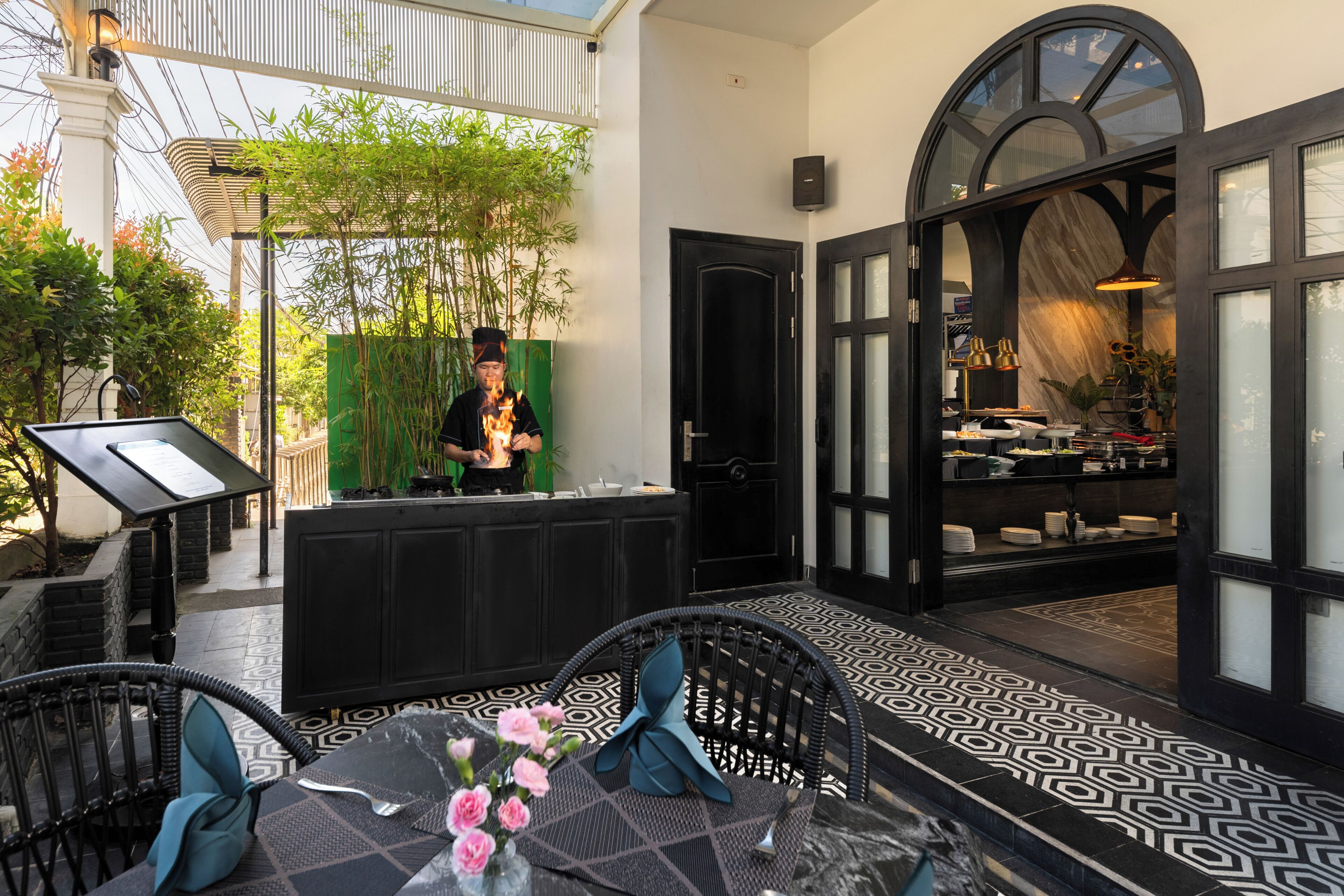 Un espace de restauration en plein air doté d’un sol à motifs noir et blanc, d’un comptoir d’accueil et offrant une vue sur l’intérieur à travers une porte vitrée.