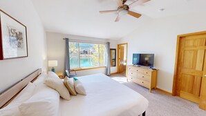 Une chambre avec un grand lit, un téléviseur posé sur une commode en bois, un ventilateur de plafond et une fenêtre donnant sur un lac.