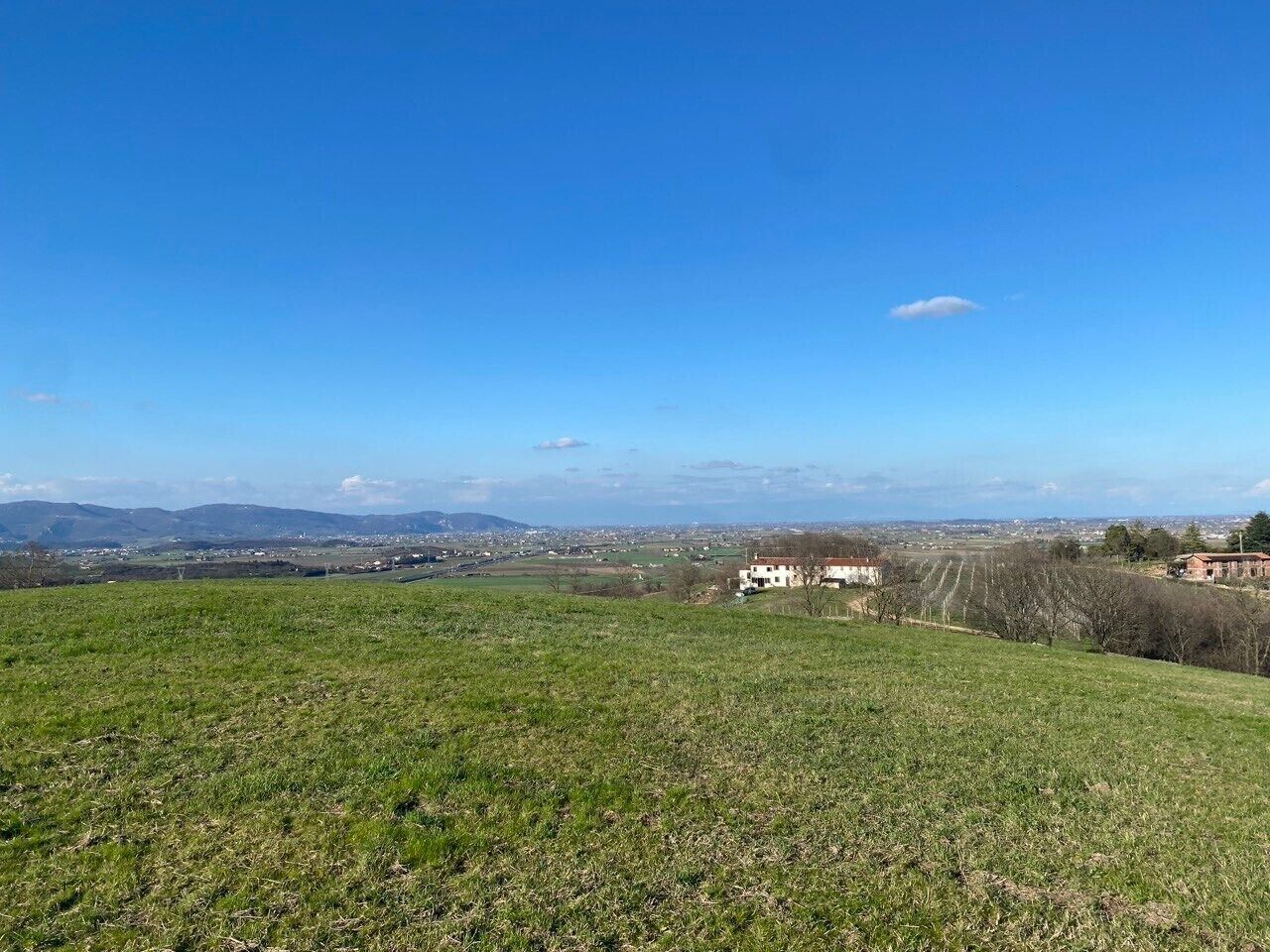 Un campo cubierto de hierba bajo un cielo despejado, con edificios lejanos y un paisaje montañoso.