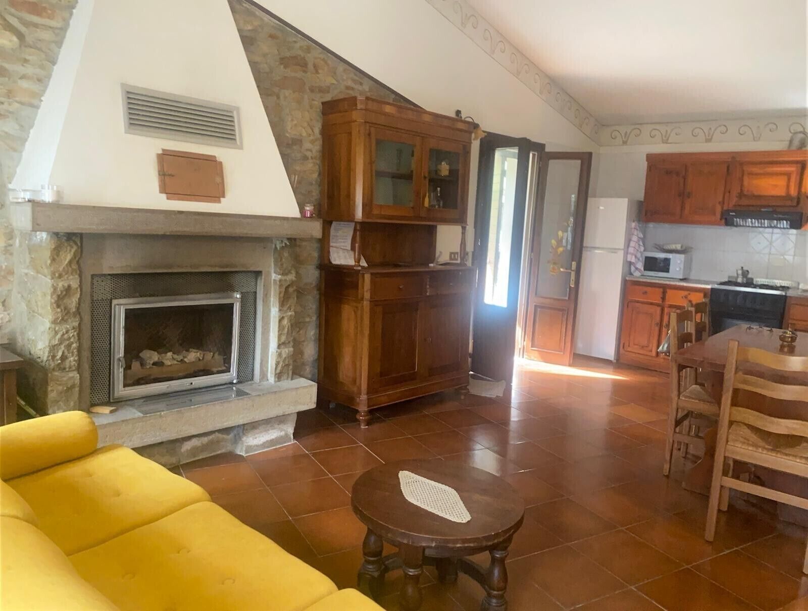 A living room with a stone fireplace, wooden cabinets, a yellow sofa, a dining table with chairs, and a kitchen area with wooden cabinets and appliances.