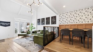 A modern living room with a green sofa, a coffee table, and a dining area with black chairs. There are large windows with a view of trees, and the walls are decorated with black and white patterned wallpaper and framed pictures.