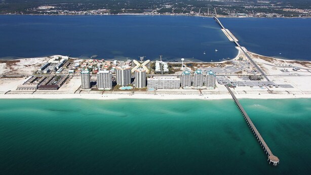 Vista aérea de una zona costera con un muelle largo que se extiende hacia el agua, rodeado de varios edificios altos, una playa y una vista despejada del océano.