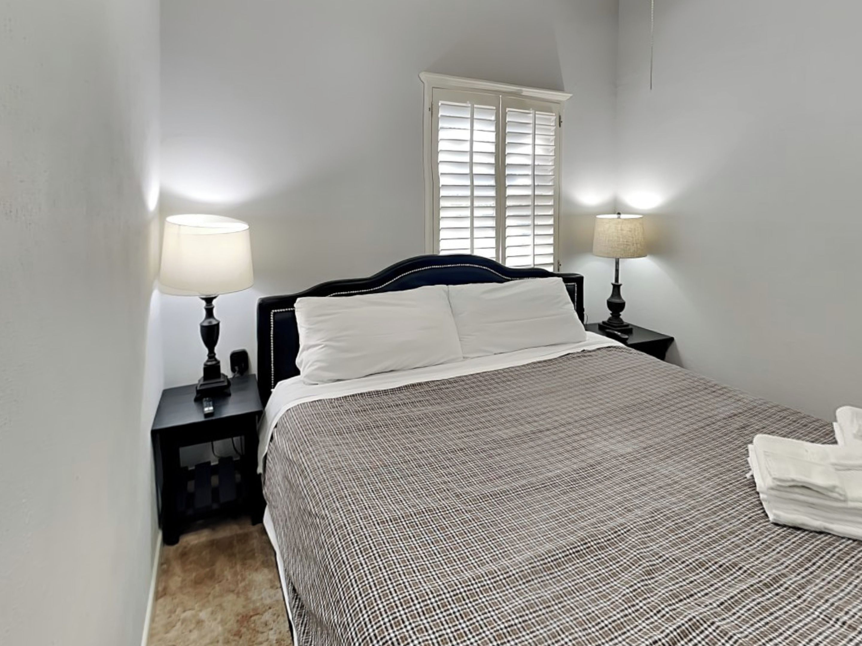 A neatly made bed with a checkered bedspread, two bedside lamps, and a window with blinds.
