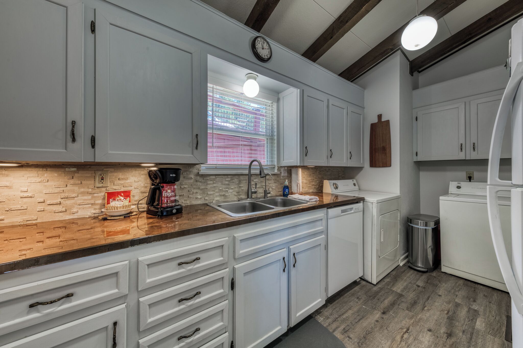 A kitchen with white cabinets, a wooden countertop, a sink, a clock, and a window with blinds.