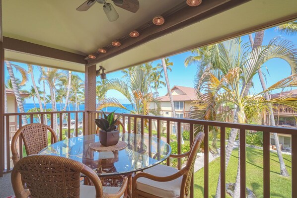 Un espacio al aire libre cubierto con una mesa de vidrio y sillas de mimbre, con vistas a un paisaje tropical con palmeras y una vista del océano.