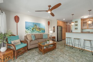 A living room with a sofa, coffee table, and bar stools. There is a ceiling fan, a refrigerator, and a kitchen area with cabinets. - KONA ISLE C38:  TOP FLOOR CORNER REMODELED W/AC & INCREDIBLE OCEAN VIEWS. (Kailua Kona)