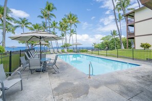 A pool area with lounge chairs and umbrellas, surrounded by palm trees and a view of the ocean.
