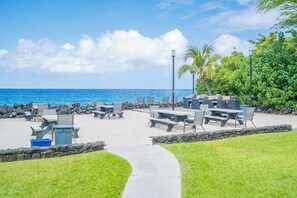 Un patio frente a la playa con mesas y sillas, un sendero y vista al océano.