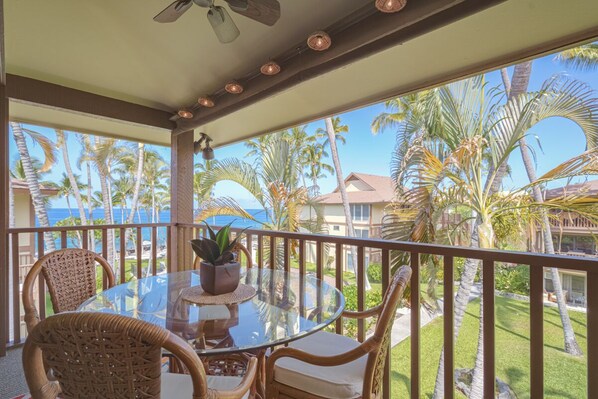 A covered outdoor area with a glass table and wicker chairs, overlooking a tropical landscape with palm trees and a view of the ocean.