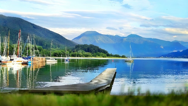 Un lac paisible, avec une jetée en bois qui s’avance sur l’eau, entouré d’une végétation luxuriante et dominé par des montagnes en arrière-plan.