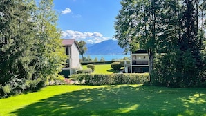 Un bâtiment à deux étages, surmonté d’un toit recouvert de tuiles rouges, entouré d’une végétation luxuriante et offrant une vue dégagée sur un lac et des montagnes en arrière-plan.