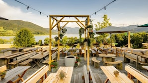 Un espace extérieur aménagé avec des tables et des bancs en bois, une structure centrale également en bois agrémentée de jardinières suspendues, et une vue sur un lac et des collines en arrière-plan.