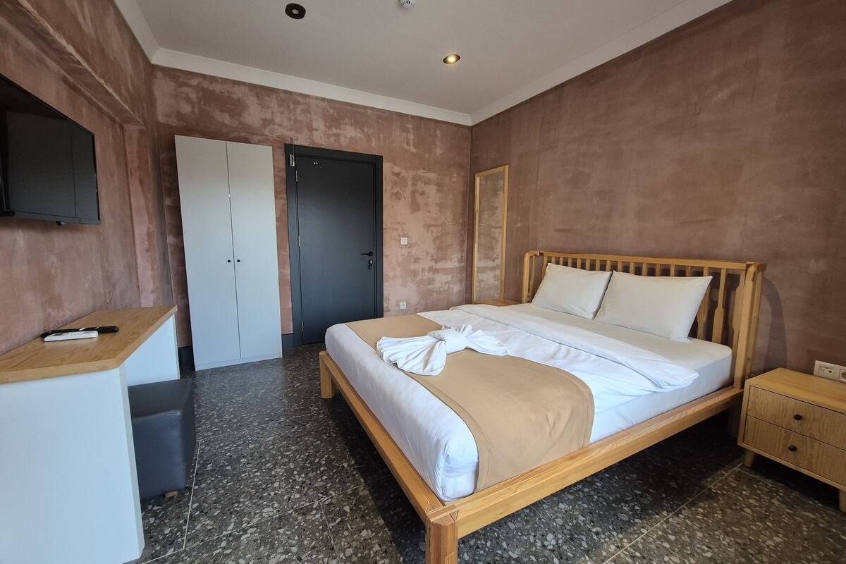 Quarto de hotel com cama, escrivaninha, guarda-roupa e televisão.