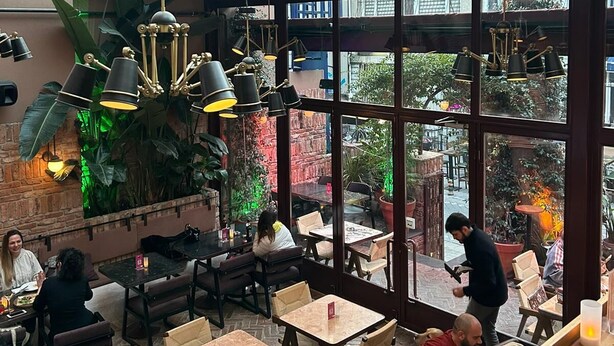 Un área de comedor interior con luz natural proveniente de amplias ventanas, mesas y sillas de madera, y lámparas colgantes.