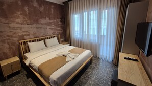 Habitación de hotel con una cama grande, una mesita de noche de madera, un televisor y una ventana con cortinas de gasa.