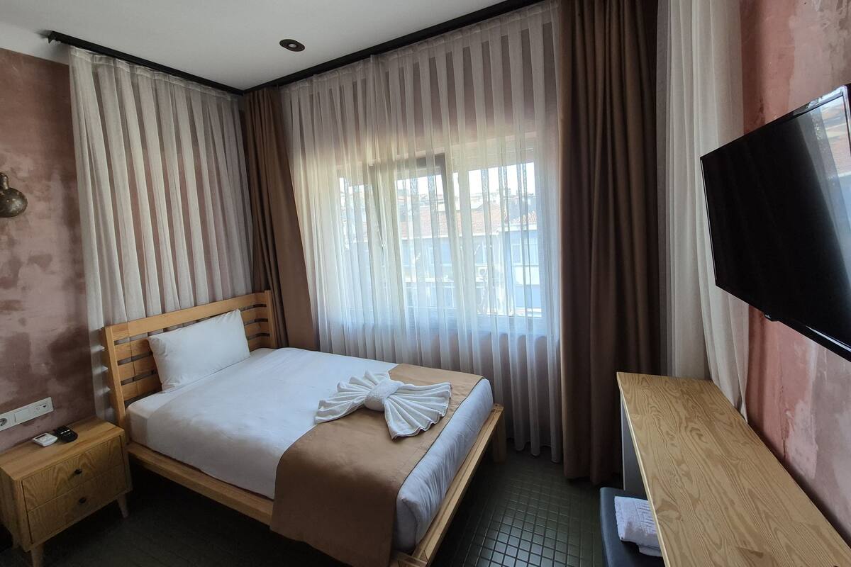 Quarto de hotel com cama, mesa de cabeceira, televisão e janela com cortinas.