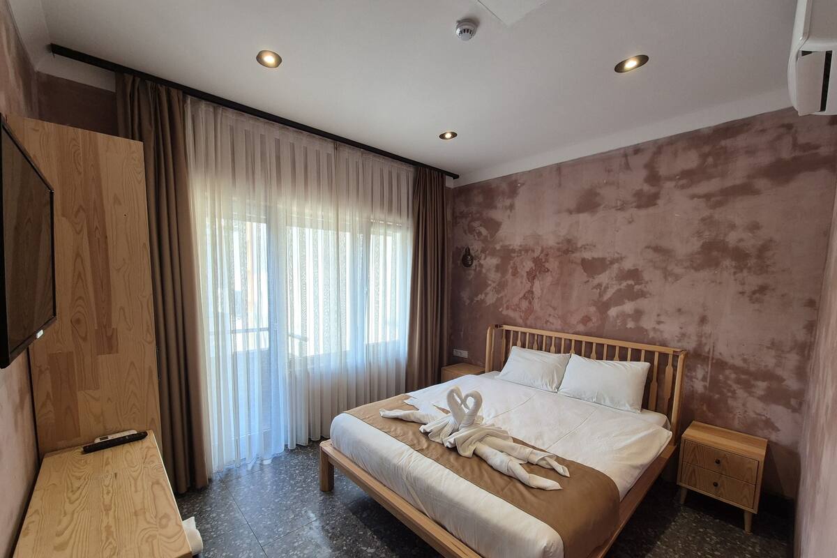 Quarto de hotel com uma cama de madeira, mesa de cabeceira, televisão e uma grande janela com cortinas.