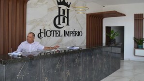 A hotel reception desk with a marble wall, a crown logo, and a receptionist. - Hotel Imperio (Aparecida do Taboado)