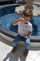 Criança sentada à beira da piscina, perto de uma fonte, vestindo camiseta branca e calças jeans.