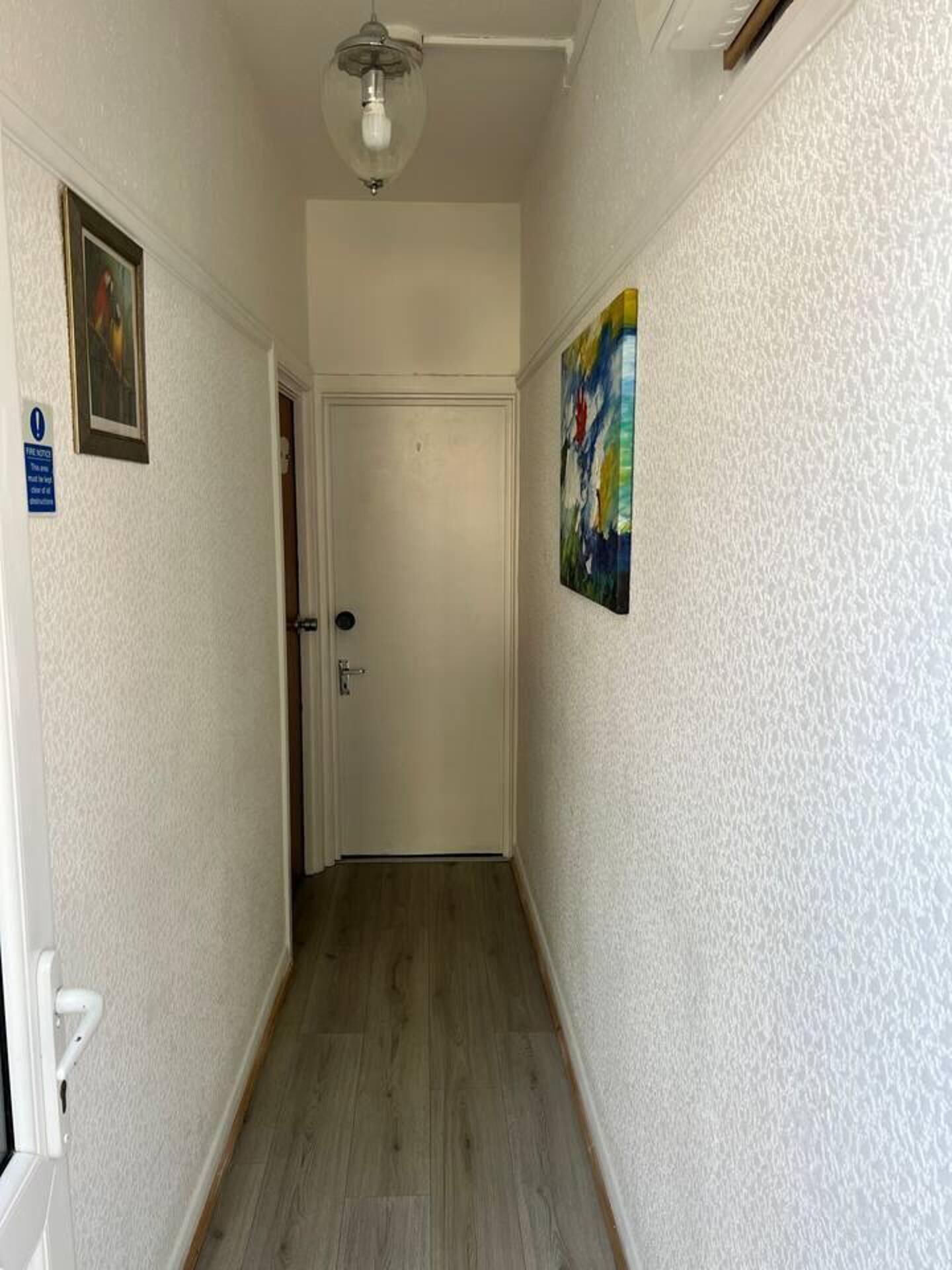 Un pasillo estrecho con suelo de madera, una puerta blanca al final y un ventilador de techo.