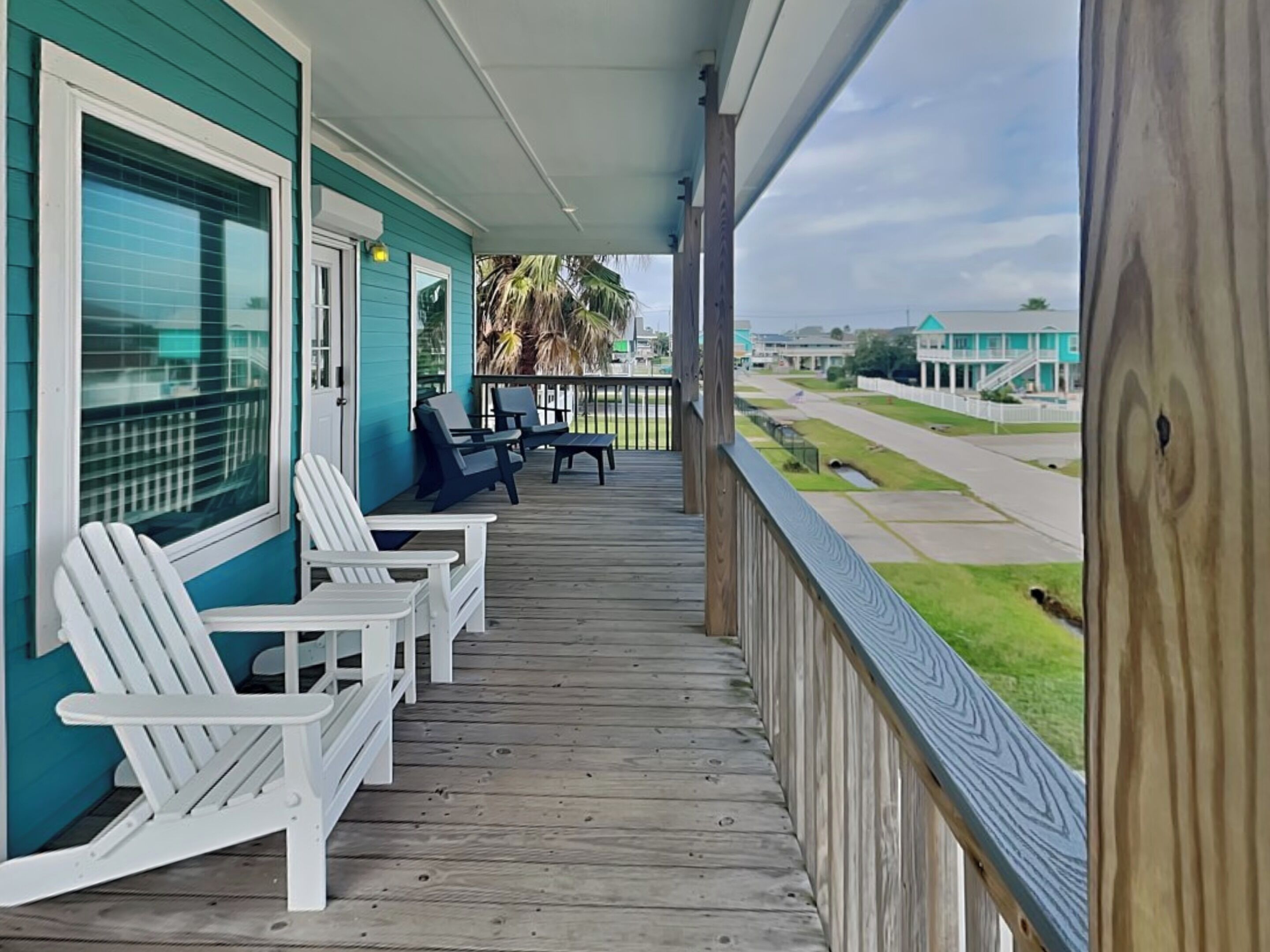 A wooden deck with white Adirondack chairs, a covered porch, and a view of a residential area with houses and a road.