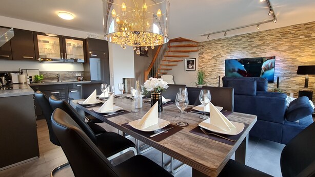 A modern dining area with a wooden table set for six, a chandelier, and a kitchen in the background.