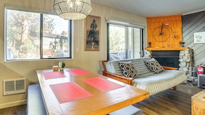 A cozy living room with a wooden dining table, a sofa with patterned cushions, a fireplace, and a large window offering a view of the outdoors. - Cozy Mammoth Lakes Studio: 5 Mi to Ski Slopes (Mammoth Lakes)