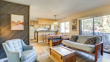 Un salon moderne avec un canapé, un fauteuil et une table basse en bois. On y trouve également un coin repas avec une table et des chaises, et une cuisine à l’arrière-plan.