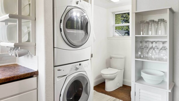 A laundry room with stacked washers and dryers, a toilet, and shelves with glassware. - Cozy Ferndale Studio on 20-Acre Working Farm! (Ferndale)