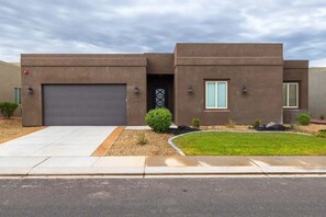 Una moderna casa a due piani con garage, un piccolo giardino anteriore con prato e arbusti, e un cielo nuvoloso.