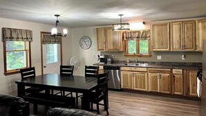 A kitchen with wooden cabinets, a dining table, and a clock on the wall. - The Hideaway: A Private Cabin in the North Maine Woods (Mount Chase)