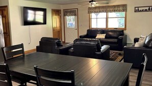 A dining area with a dark wooden table and chairs, a black leather sofa, and a ceiling fan. - The Hideaway: A Private Cabin in the North Maine Woods (Mount Chase)