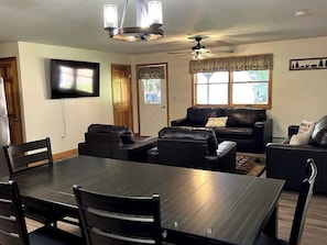 A dining area with a dark wooden table and chairs, a black leather sofa, and a ceiling fan. - The Hideaway: A Private Cabin in the North Maine Woods (Mount Chase)