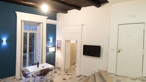 Une pièce avec un mur bleu, une porte blanche, un téléviseur fixé au mur, une table avec une chaise, un canapé et un escalier.